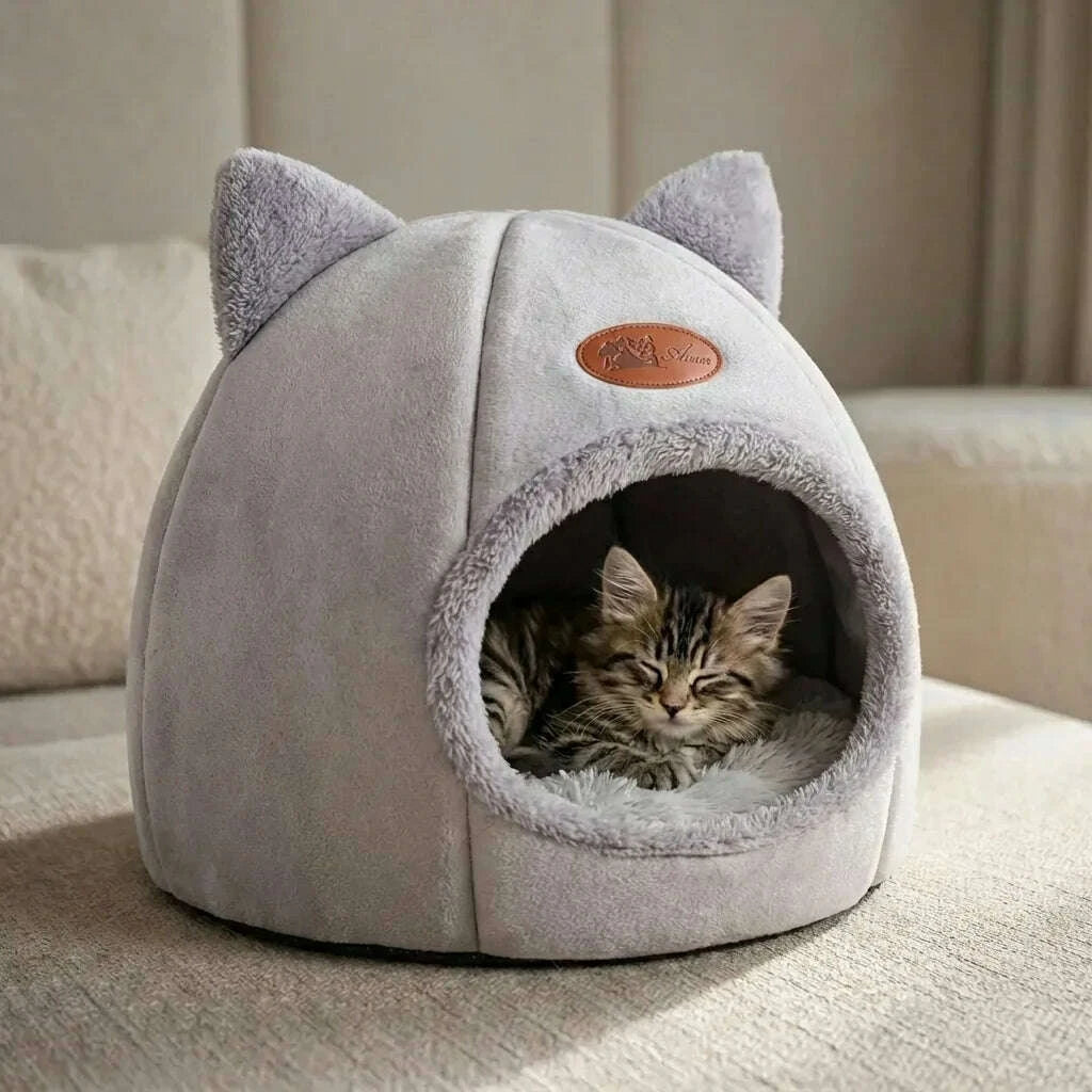 Chaton tigré endormi dans un abri douillet gris à oreilles de chat, posé sur un canapé beige