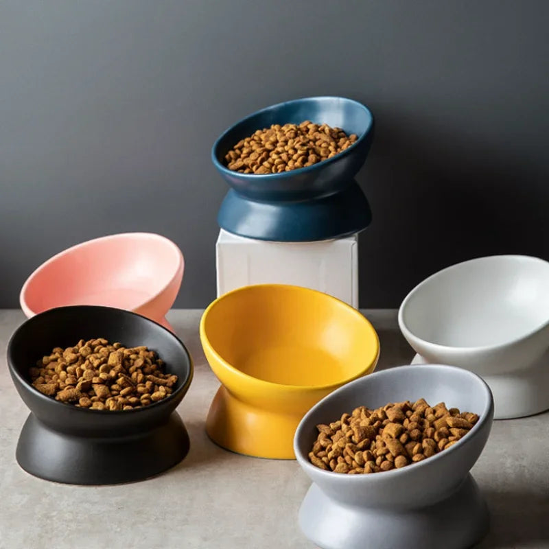Five tilted ceramic cat bowls in black, pink, blue, yellow, and gray, filled with kibble