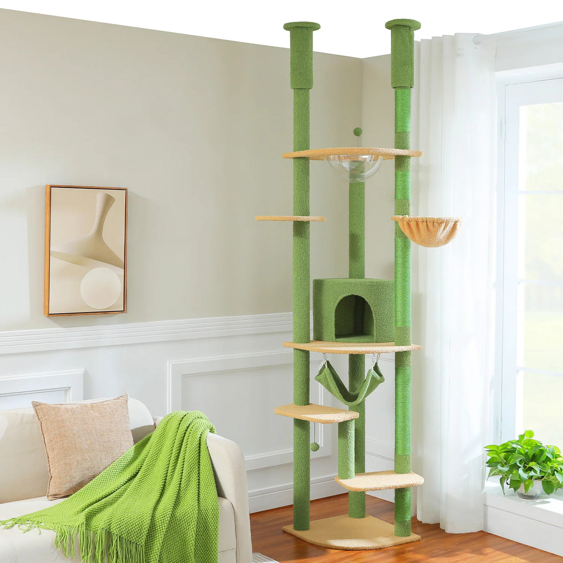 Arbre à chat vert et beige vu de face dans un séjour clair, avec niche, hamac vert, panier et bulle transparente