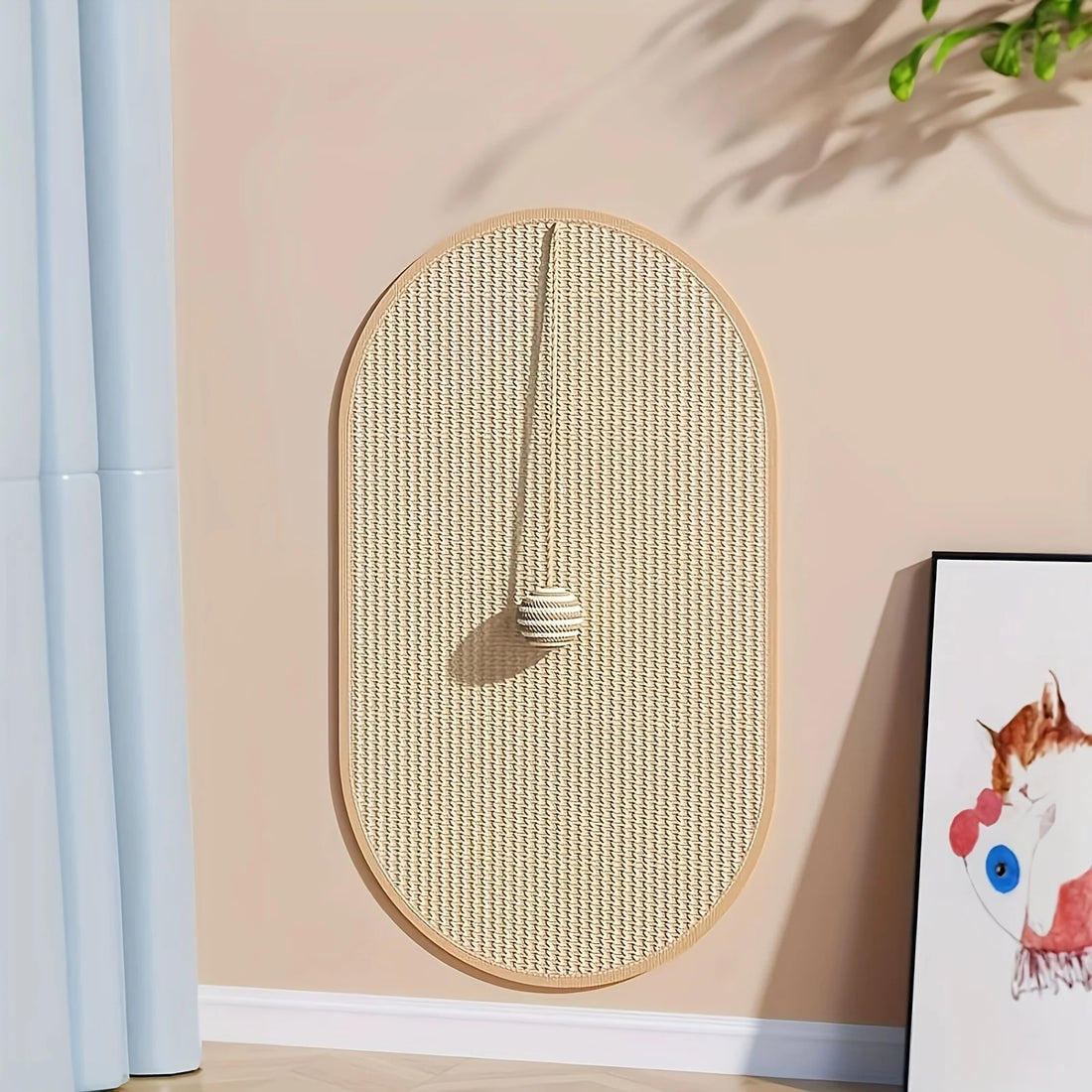 Griffoir mural ovale en sisal fixé au mur beige, balle rayée suspendue, à côté d'un rideau bleu clair