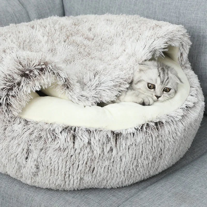 Chaton gris qui regarde sous la capuche rabattue d'un couchage en peluche taupe posé sur un canapé