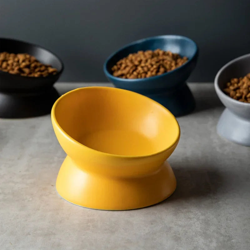 Empty yellow ceramic cat bowl in the foreground, raised base and tilt clearly visible, blurred background