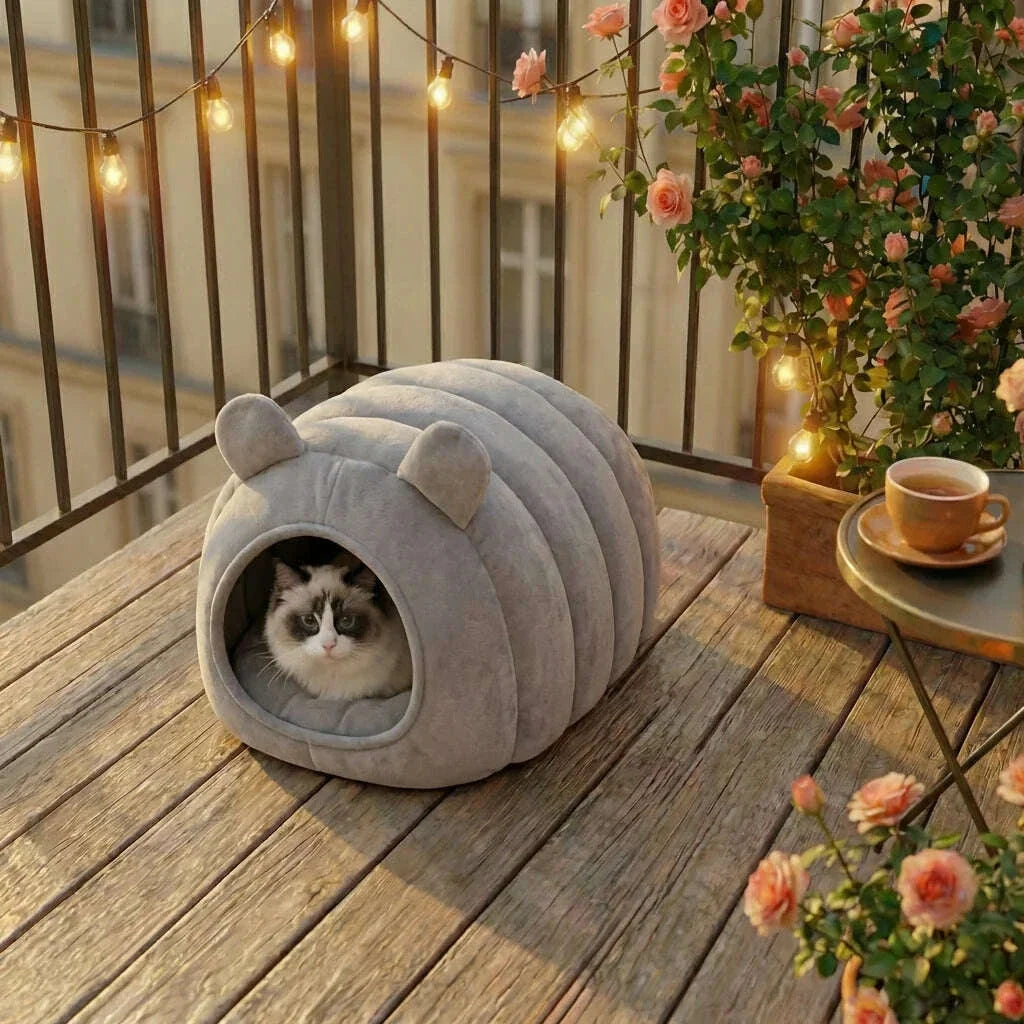 Chat ragdoll dans une niche douillette grise à oreilles posée sur un balcon en bois fleuri, guirlande lumineuse
