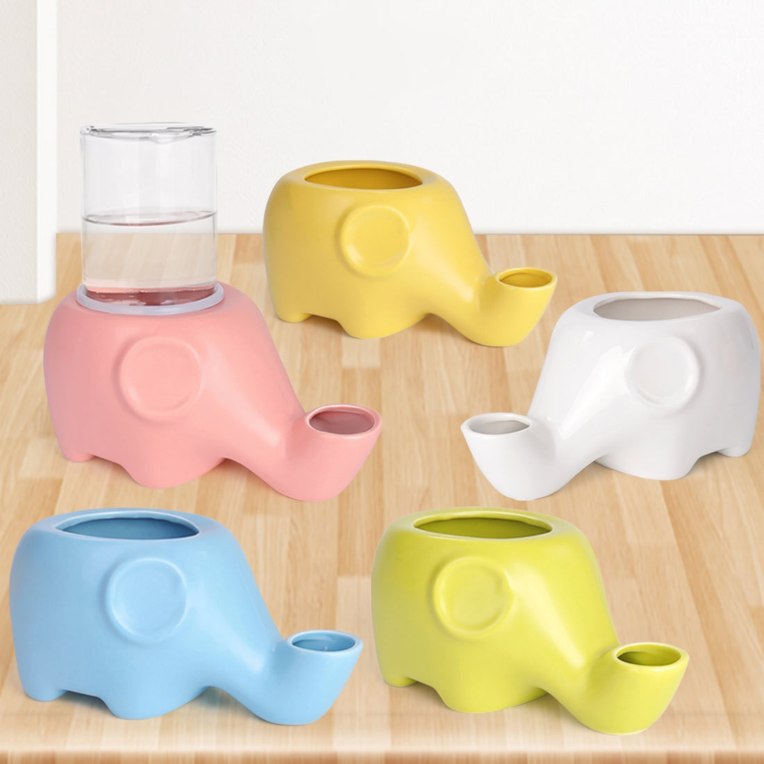 Abreuvoirs éléphant en céramique de cinq coloris posés sur un parquet clair, réservoir en verre sur le modèle rose