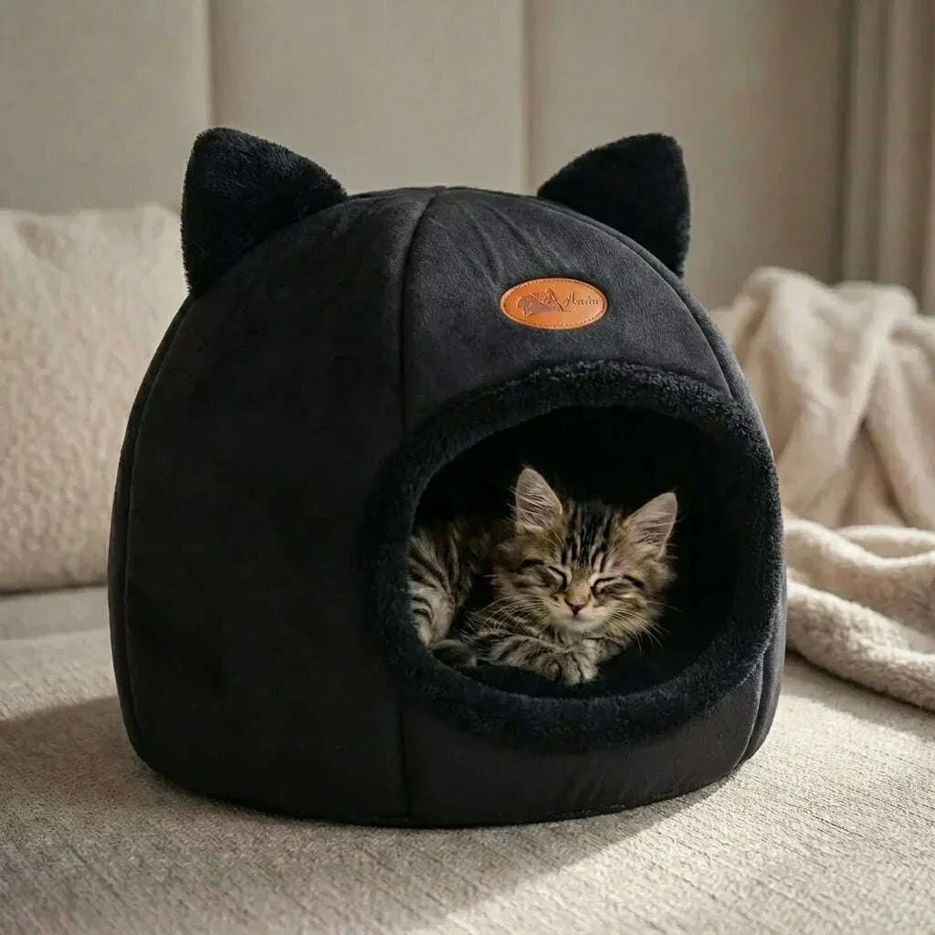 Petit chat tigré endormi dans une cabane noire à oreilles, sur un canapé avec plaid crème