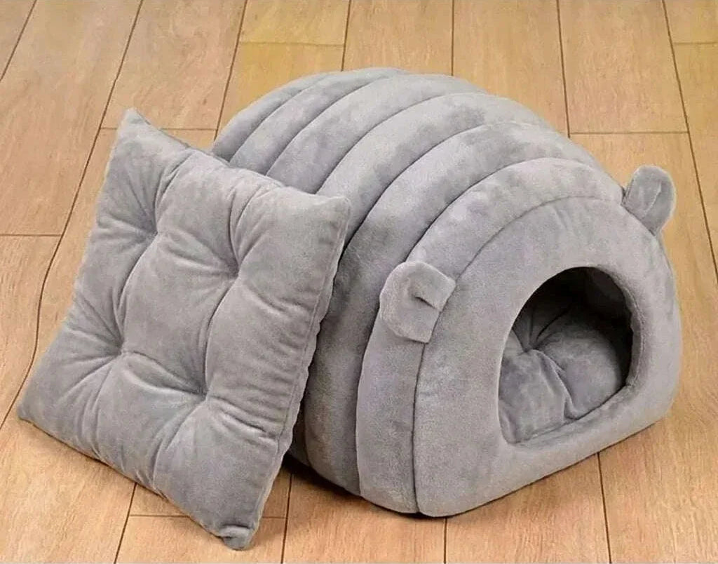 Abri en peluche grise pour chat avec son coussin matelassé carré posé à côté sur un parquet clair