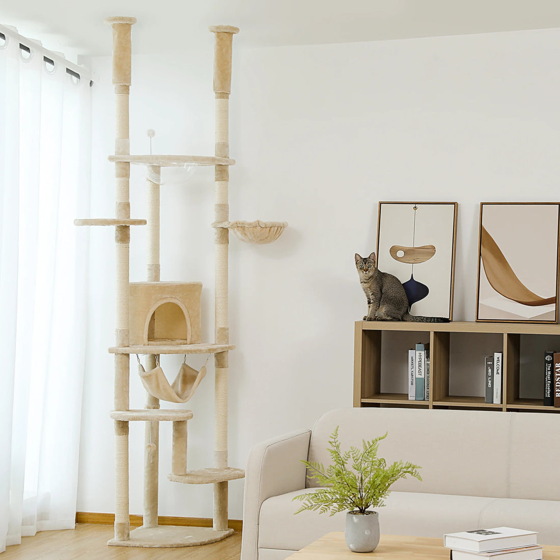 Arbre à chat beige très haut près d'un canapé, avec un chat tigré assis sur l'étagère voisine
