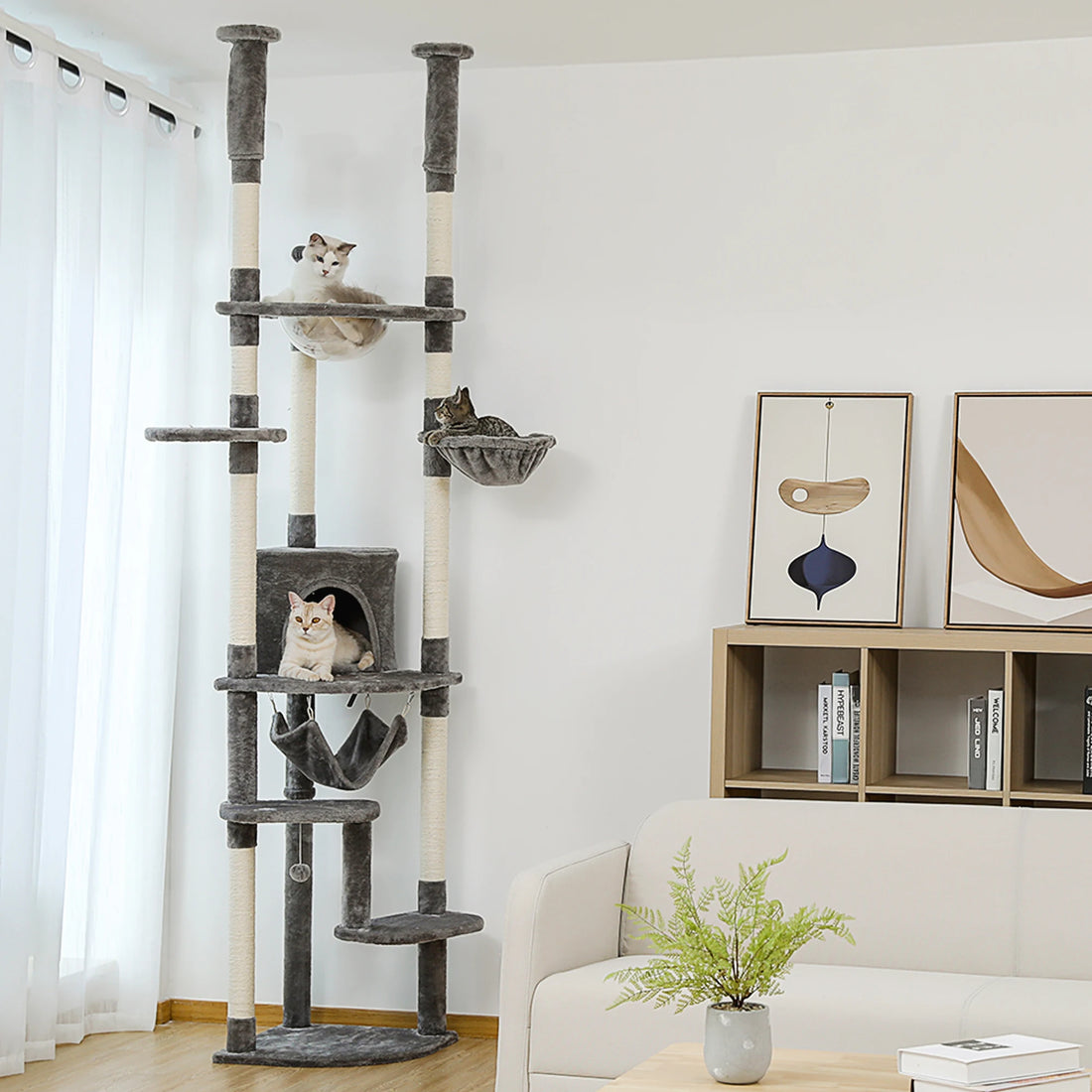 Arbre à chat gris dans un salon avec un chat blanc en haut, un tigré dans le panier et un chat clair dans la niche