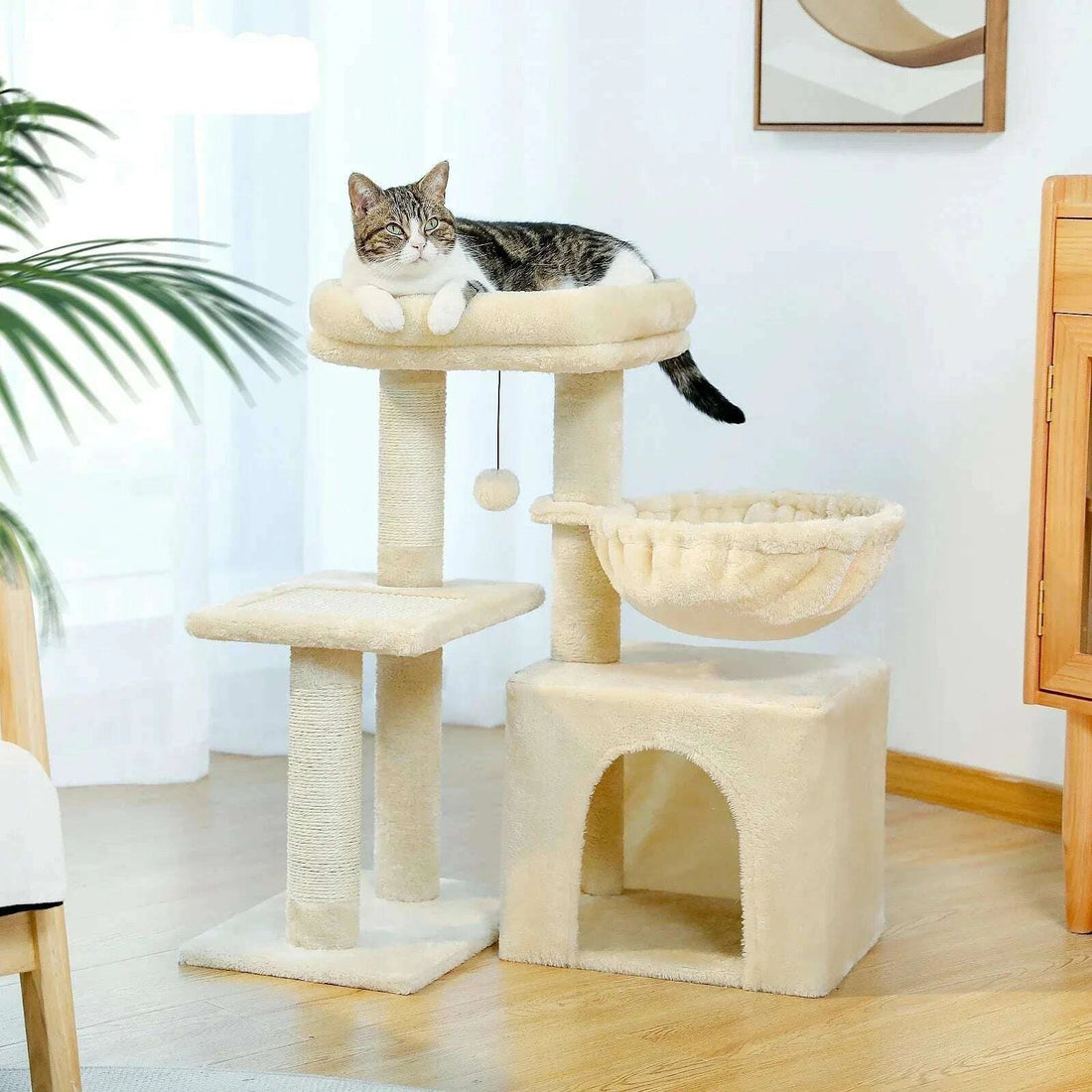 Meuble à chat beige dans un salon lumineux, chat tigré couché sur le plateau du haut, plante verte à gauche