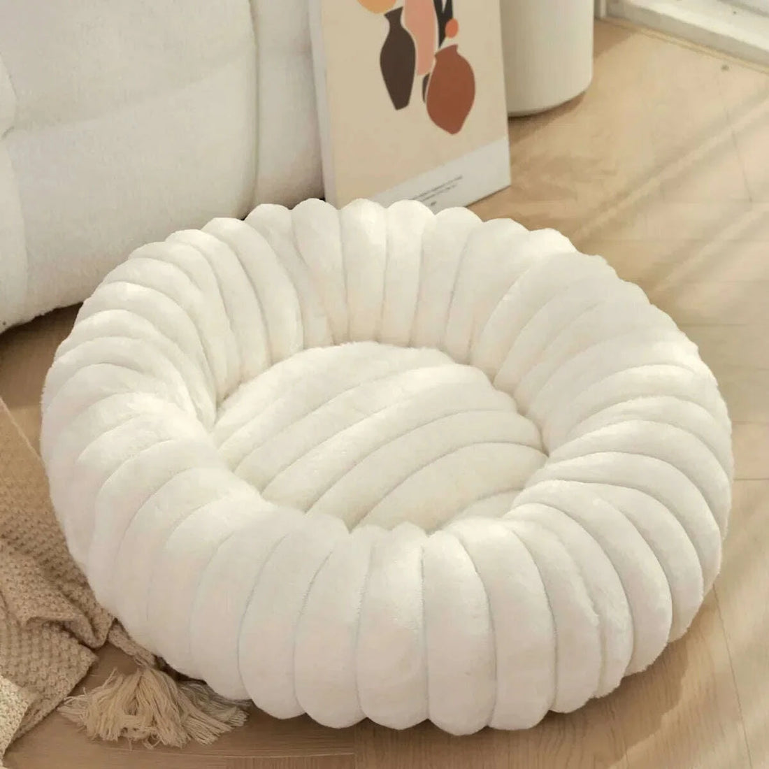 Lit douillet blanc en forme de beignet pour chat, à côté d'un canapé crème et d'un plaid à franges