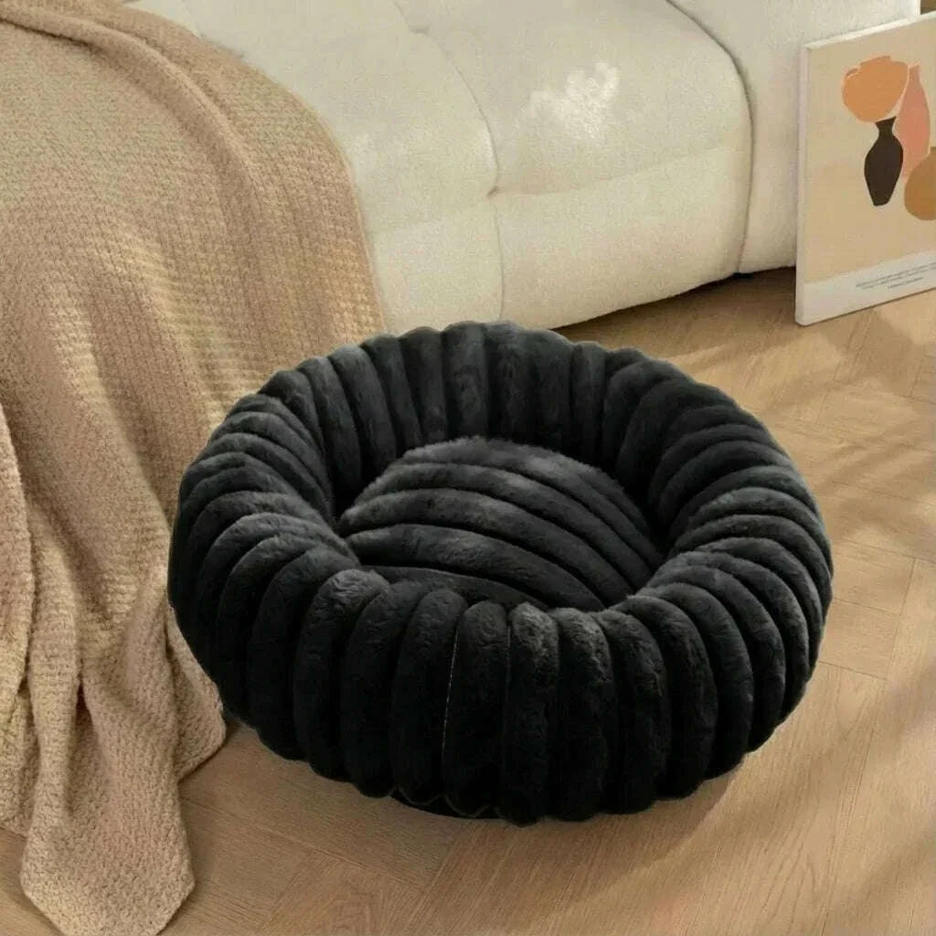 Couchage rond noir en peluche à rebord matelassé, installé sur le parquet devant un canapé blanc et un plaid beige