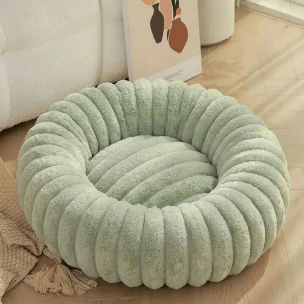 Corbeille ronde en fausse fourrure vert sauge vide, posée au sol près d'un canapé blanc et d'un plaid à franges