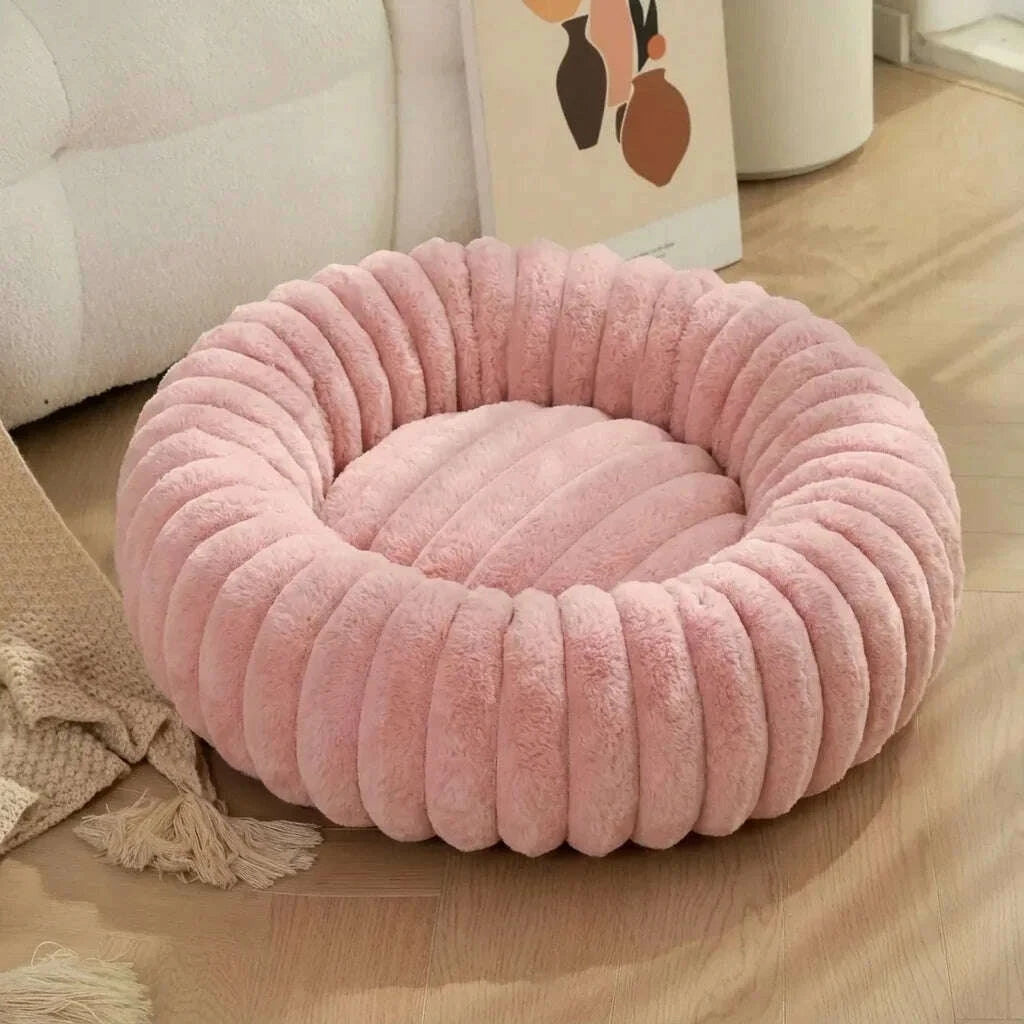 Nid rond en fausse fourrure rose pâle sur un parquet clair, à côté d'un canapé blanc bouclé et d'un plaid à franges